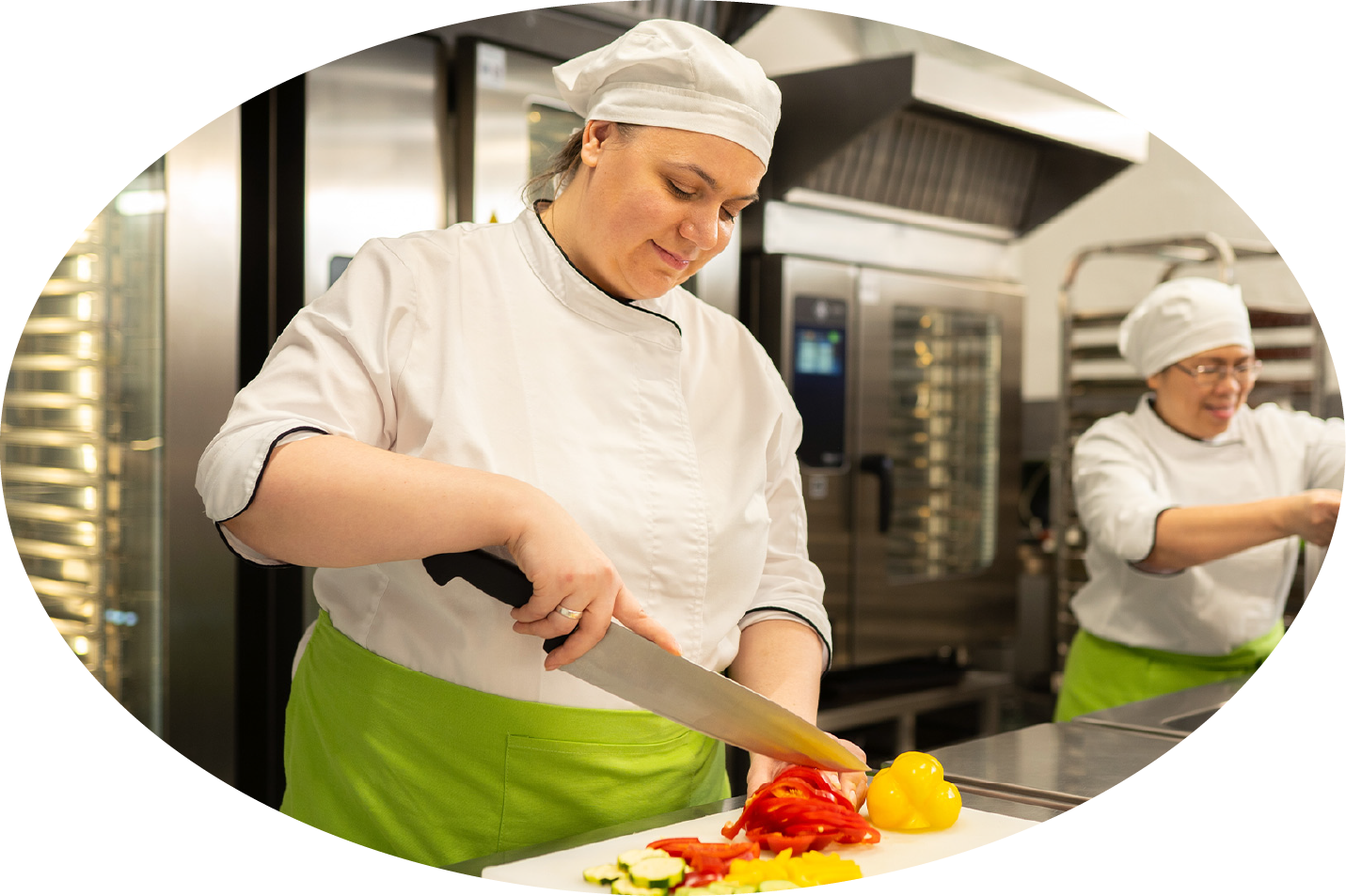 Zwei Köchinnen in weißer Arbeitskleidung und grünen Schürzen in einer Küche.